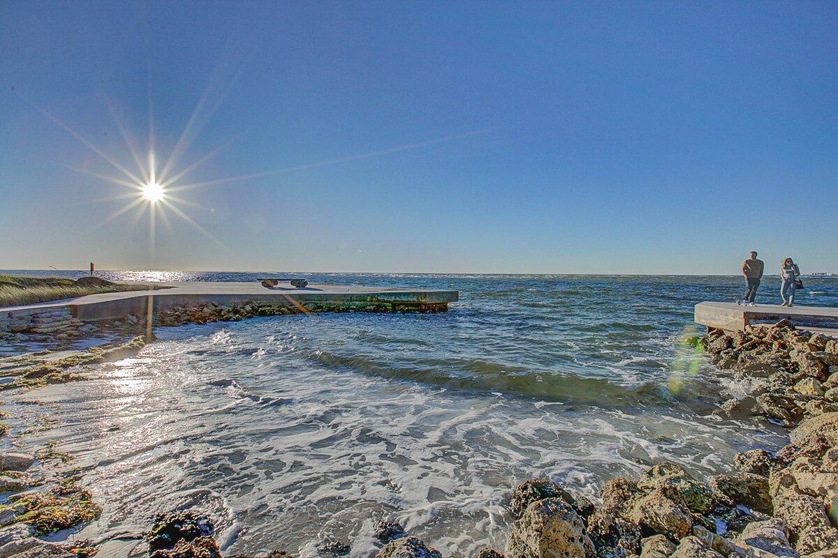 A coastal scene with a pier extending into the water, a clear sky, and two individuals standing on the pier.