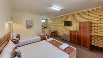A hotel room with two beds, a kitchenette, a dining table with chairs, a TV mounted on the brick wall, and a wooden wardrobe.