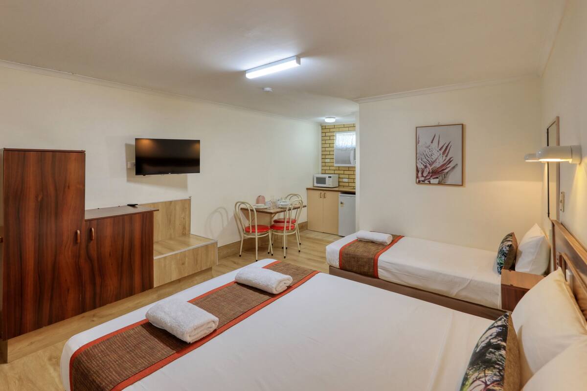 A hotel room with two beds, a wooden wardrobe, a television, a kitchenette, and a dining area.