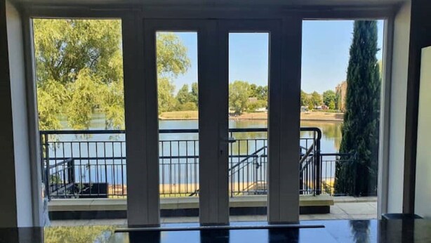 A view through a glass door with a metal railing, leading to a balcony overlooking a body of water and greenery. - Executive Lakeside 4 Bed Townhouse (Milton Keynes)