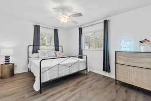 A bedroom with a bed, a ceiling fan, a bedside table with a lamp, a dresser, and a window with curtains.