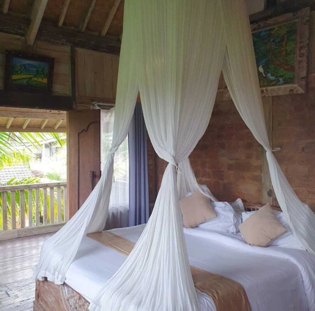 A room with a bed under a canopy, a wooden ceiling, and a view of greenery outside.