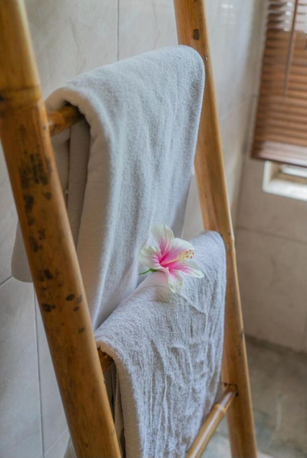 A wooden chair with a white towel draped over it, a pink and white flower, and a window with blinds in the background.