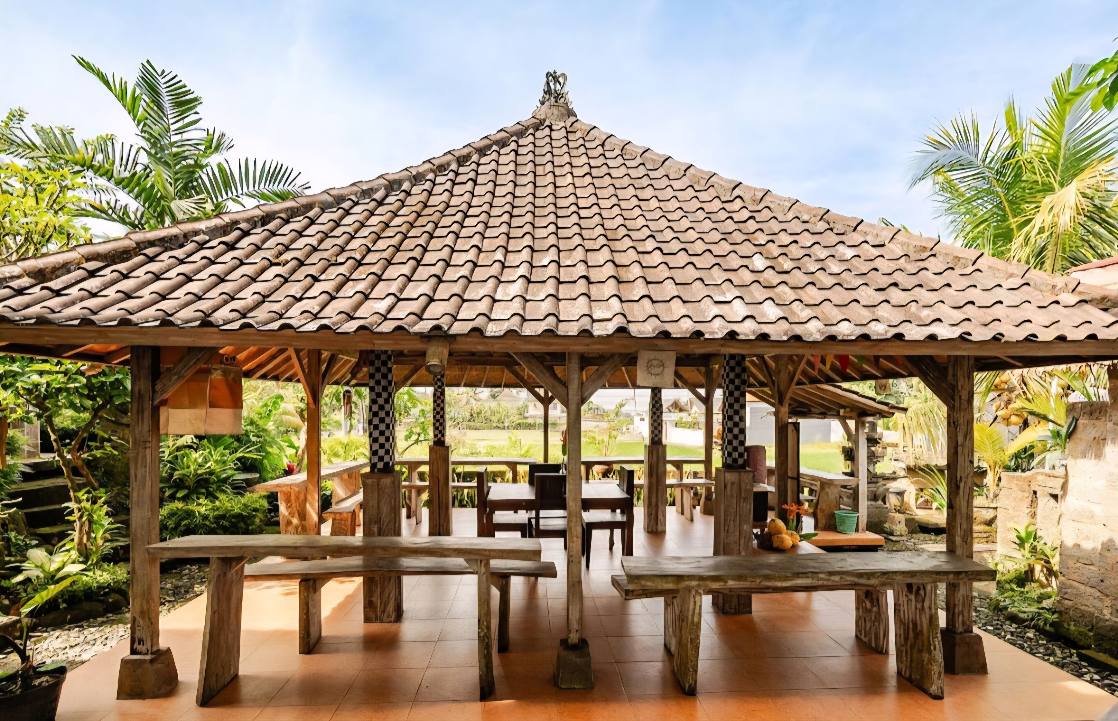 A gazebo with a tiled roof, wooden benches, and tables, surrounded by greenery and palm trees.
