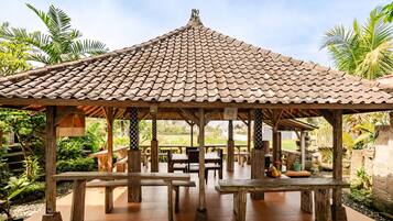 A gazebo with a tiled roof, wooden benches, and tables, surrounded by greenery and palm trees.