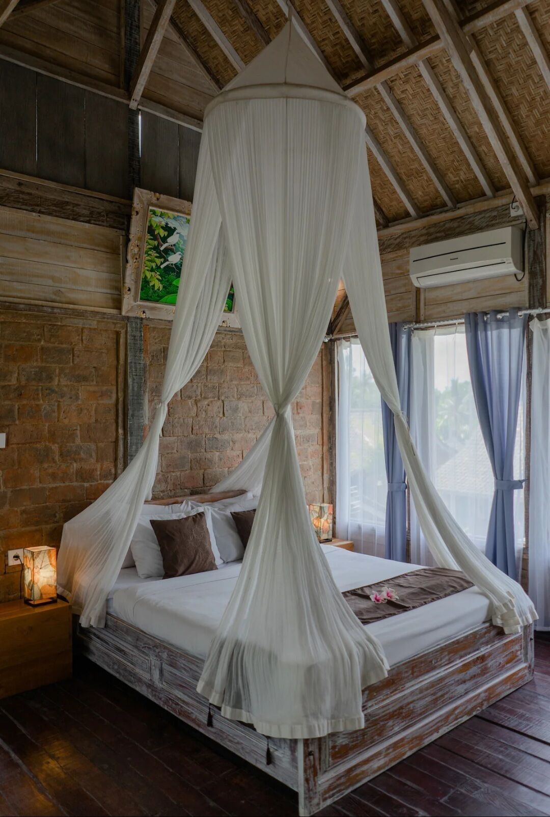 Una habitación con cama bajo un dosel, paredes de ladrillo, un equipo de aire acondicionado y una ventana con cortinas.