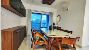 Un comedor con mesa de vidrio, sillas de madera, un armario de madera, televisor montado en la pared, ventana con cortinas y un ventilador de techo.