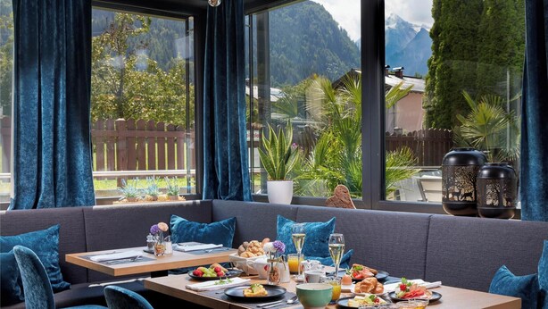 Un comedor con vistas a las montañas, una mesa de madera dispuesta para el desayuno y asientos tapizados en azul.