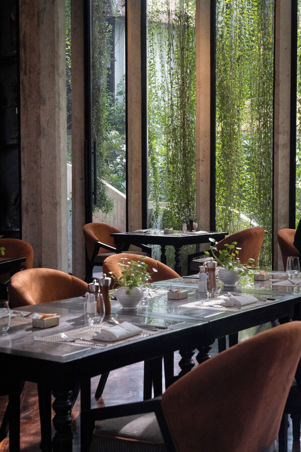 A dining area with a long table set for four, surrounded by chairs, with a view of greenery through large windows. - Hotel Vellita Siem Reap (Siem Reap)