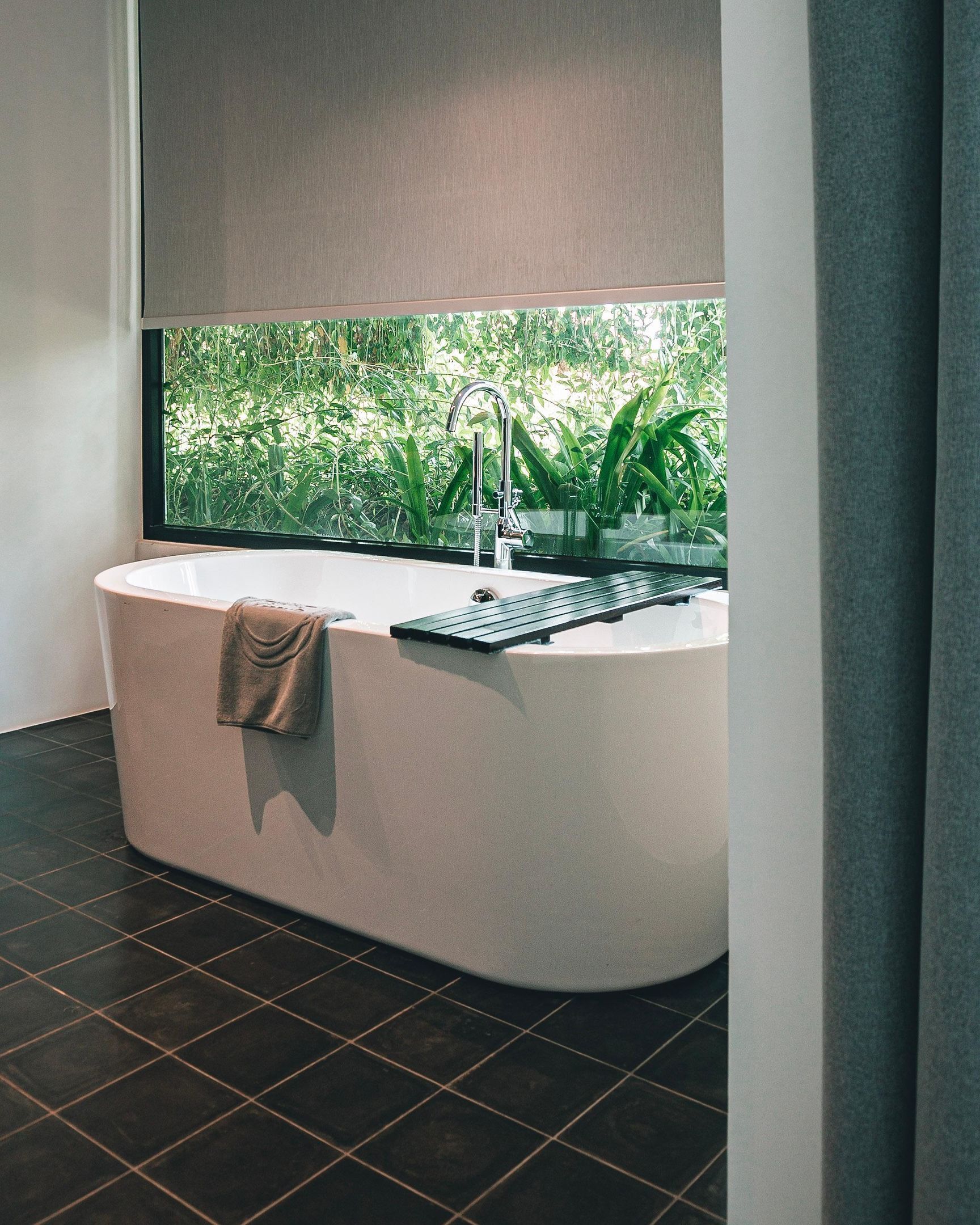 A modern bathroom with a freestanding bathtub, a window with a view of greenery, and dark tiled flooring.