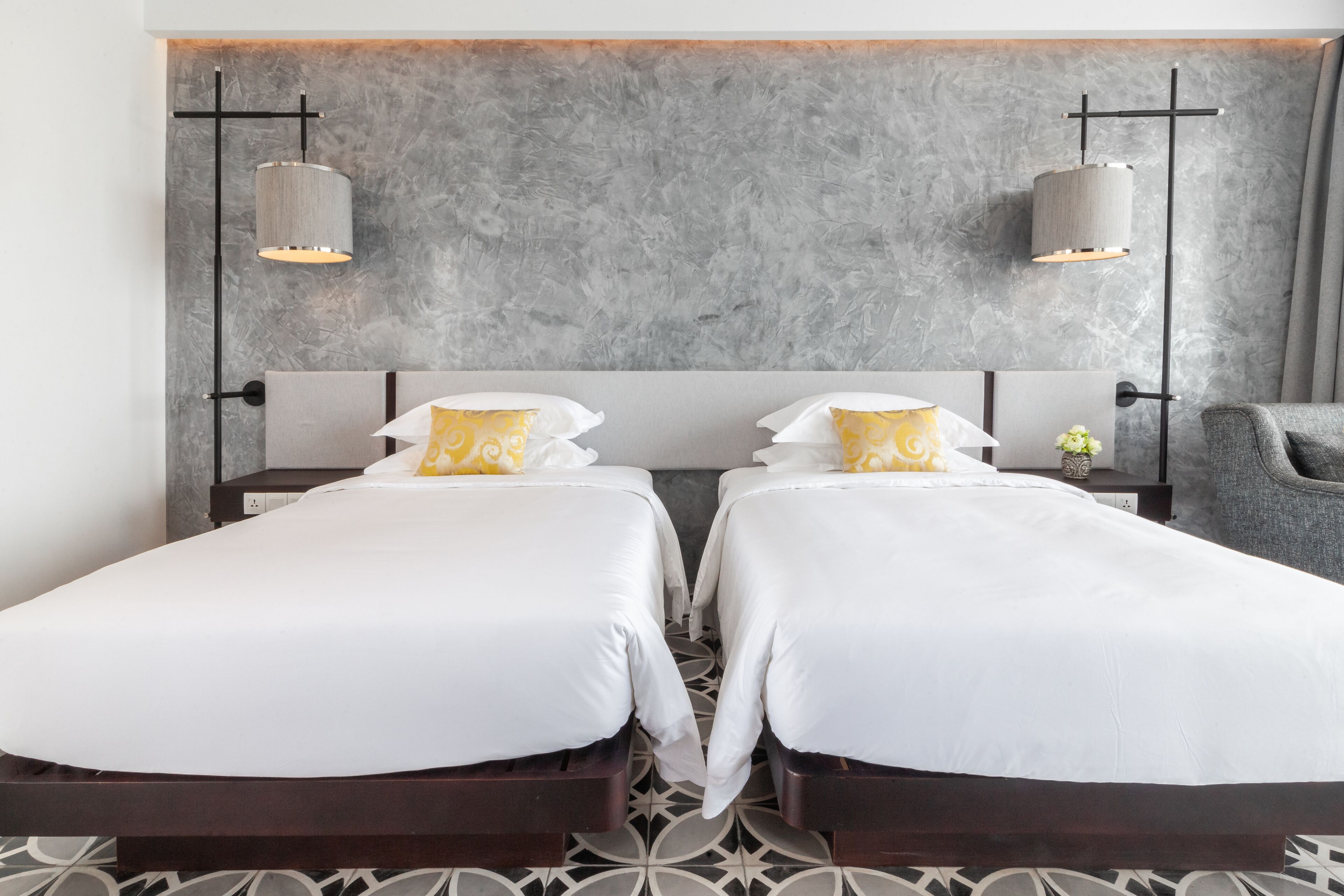 A hotel room with two beds, a grey textured wall, and two pendant lights.