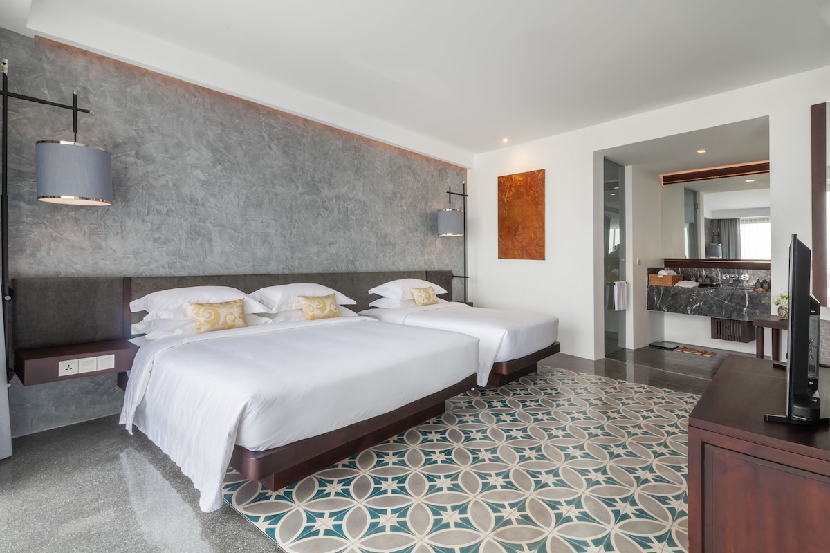 Een moderne hotelkamer met twee bedden, een gestructureerde grijze muur, een vloer met patroon, en een badkamer zichtbaar door een geopende deur.