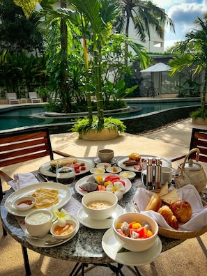 A table set with breakfast items, including fruits, bread, and beverages, in a tropical outdoor setting with a pool and lush greenery. - Hotel Vellita Siem Reap (Siem Reap)