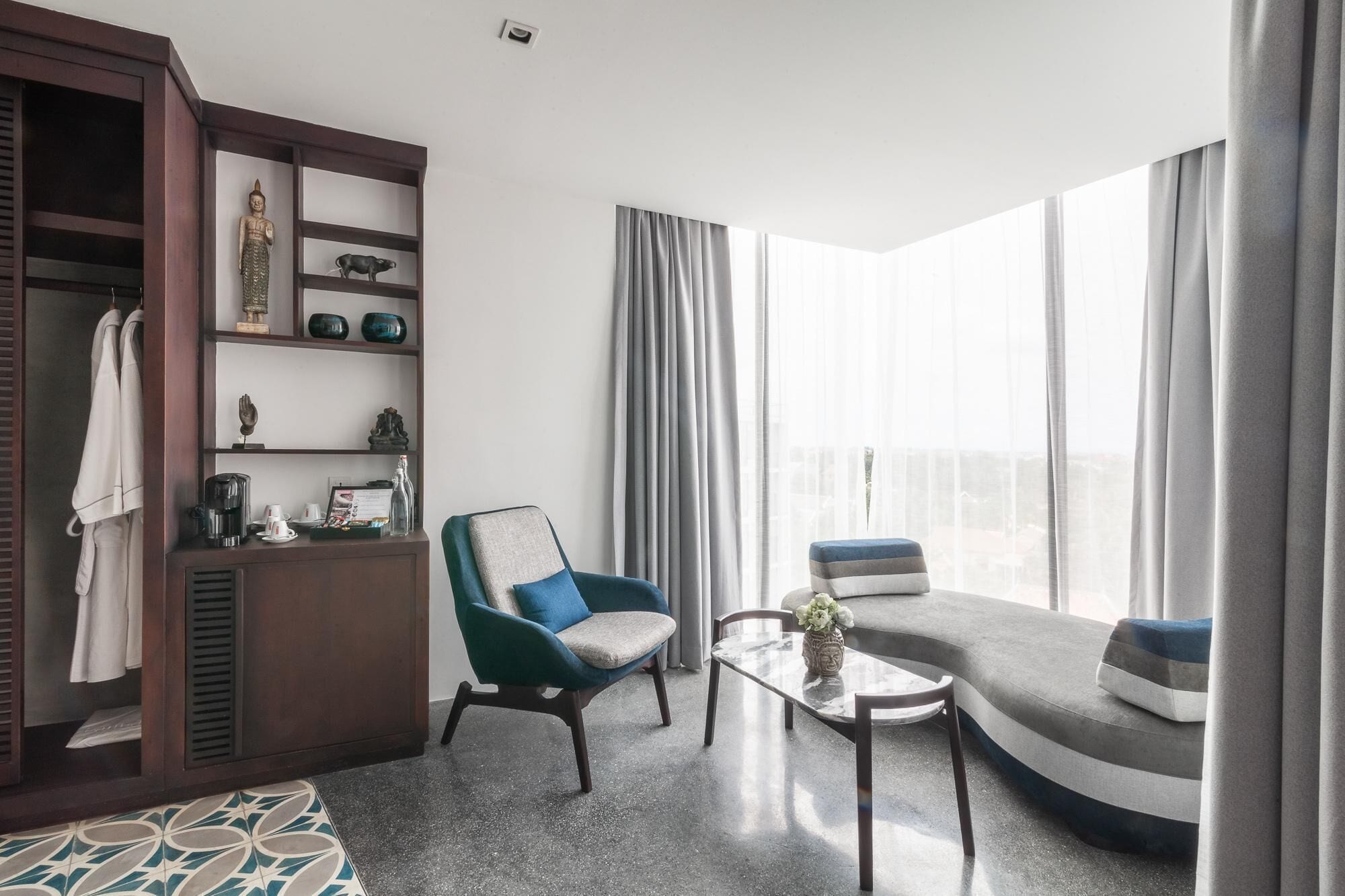 Un salon moderne avec un canapé, un fauteuil et une table basse. On y trouve un meuble en bois avec des étagères et une statue. La pièce est dotée de grandes fenêtres avec des rideaux.