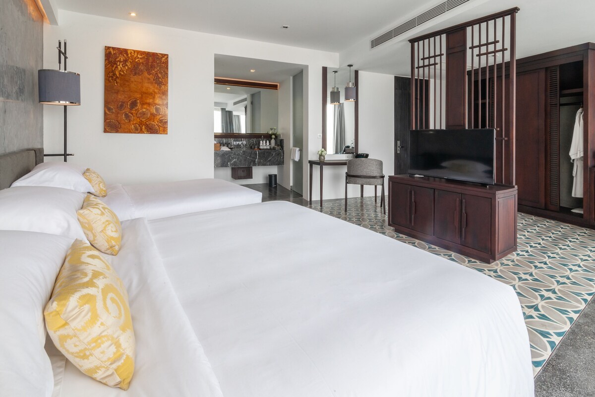 Habitación de hotel con dos camas, televisión, escritorio y baño visible a través de una puerta abierta.