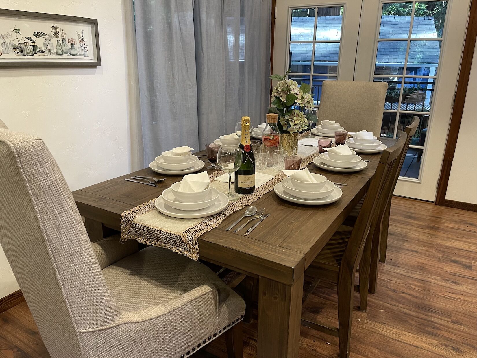 Une table à manger dressée avec de l’assiette blanche, des verres et des couverts, une bouteille de vin et un vase rempli de fleurs.