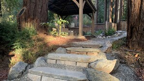 A stone pathway leading to a wooden gazebo surrounded by large trees and greenery.