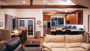 A spacious living room with a leather sofa, wooden dining table, and a kitchen area with wooden cabinets and a large window.