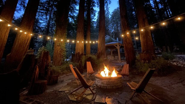 A fire pit surrounded by wooden chairs in a forested area with string lights.