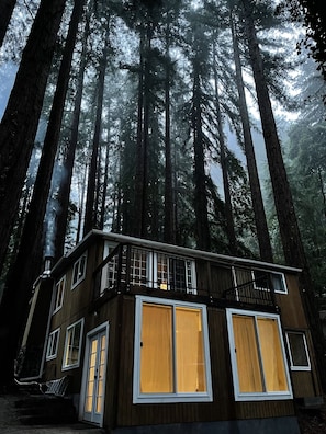 A cabin with a balcony, large windows, and a chimney emitting smoke, surrounded by tall trees.