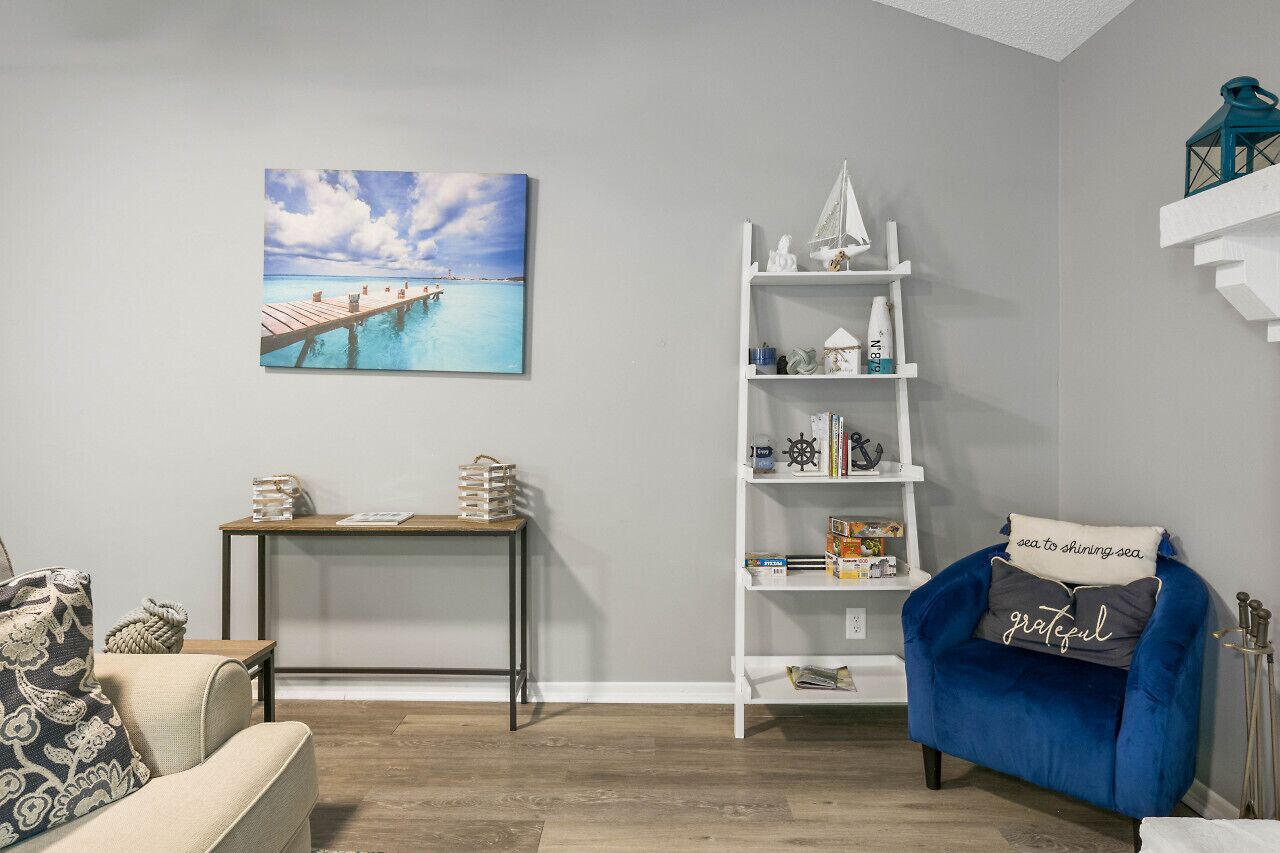 A living room with a blue armchair, a white bookshelf, a wooden side table, and a painting of a pier.