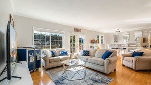 A spacious living room with two sofas, a coffee table, and a TV. There are large windows and a view of the outdoors.