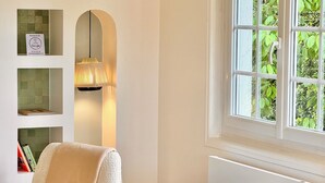 A cozy room with a bed, a chair, a shelf with books, a lamp, and a window with greenery outside.