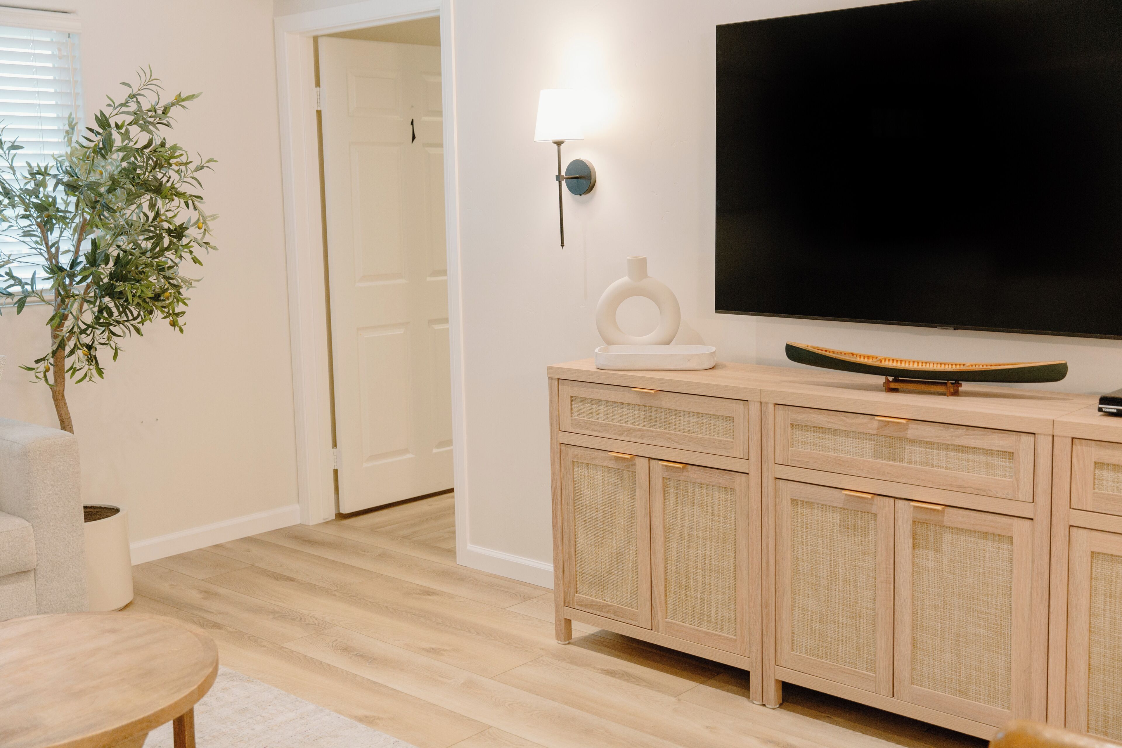 En stue med et fladskærms-tv, et fletskab, et rundt bord og en potteplante.
