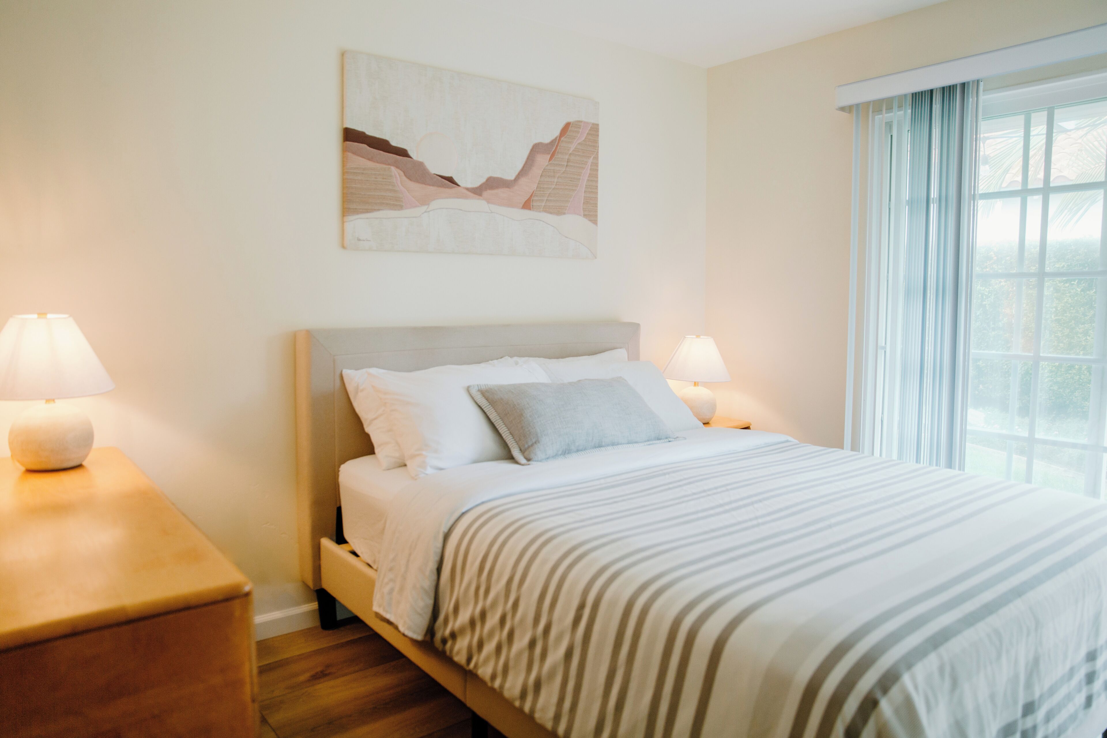 A bedroom with a bed, two bedside tables, a window with curtains, and a wall-mounted artwork.