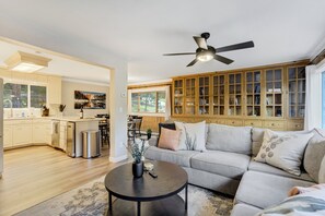 Eine moderne Küche mit Holzschränken, ein Essbereich mit Tisch und Stühlen sowie ein Wohnzimmer mit Sofa und Deckenventilator.
