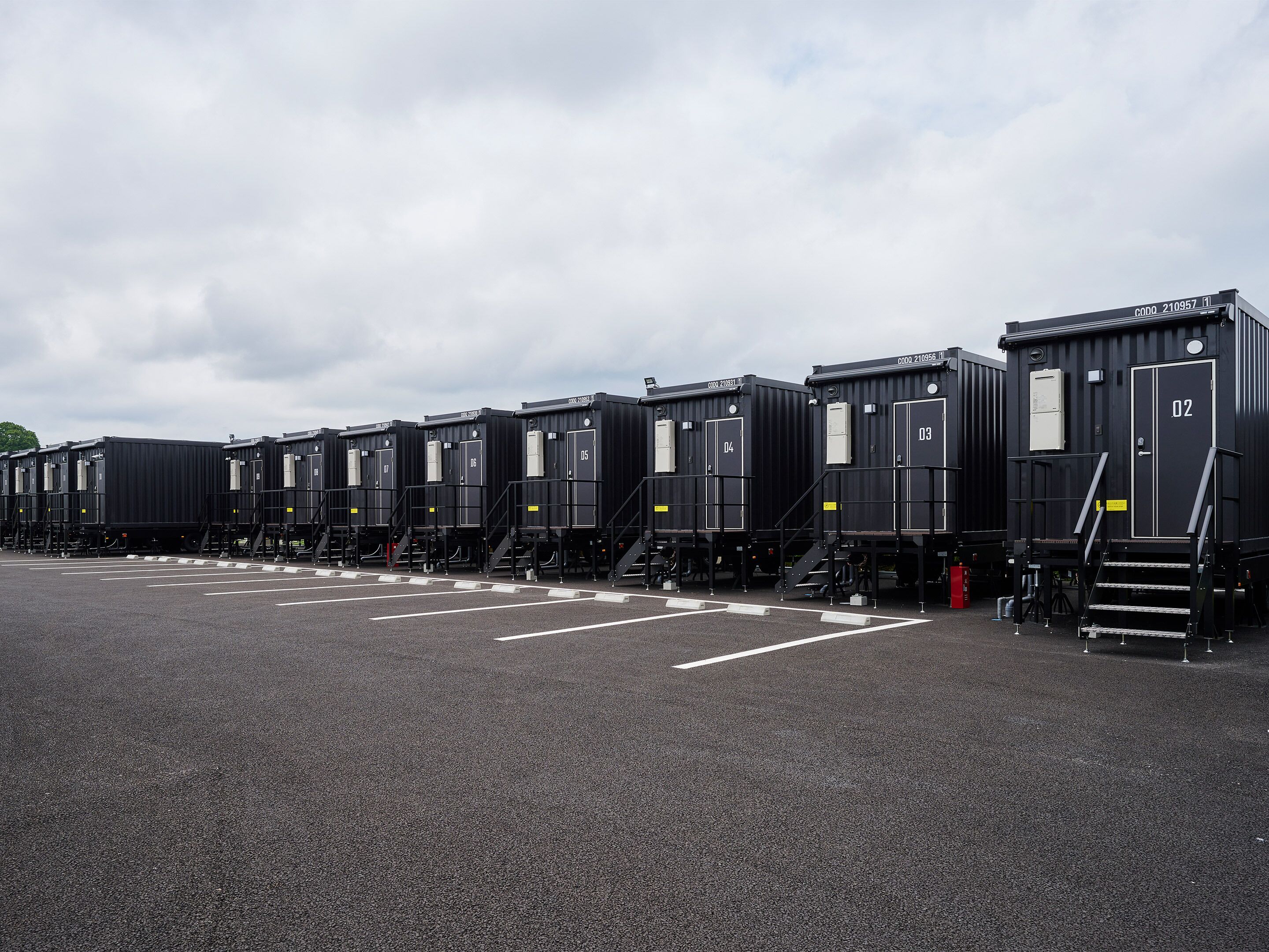 Een rij zwarte, industrieel ogende treinwagons, genummerd van 02 tot 07, geparkeerd op een terrein met witte parkeerlijnen.