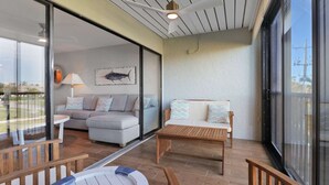A spacious balcony with a sliding glass door leading to a living area, furnished with a sofa, a wooden table, and a boat-shaped chair. - Cozy 1-bedroom condo with pool access (Marco Island)