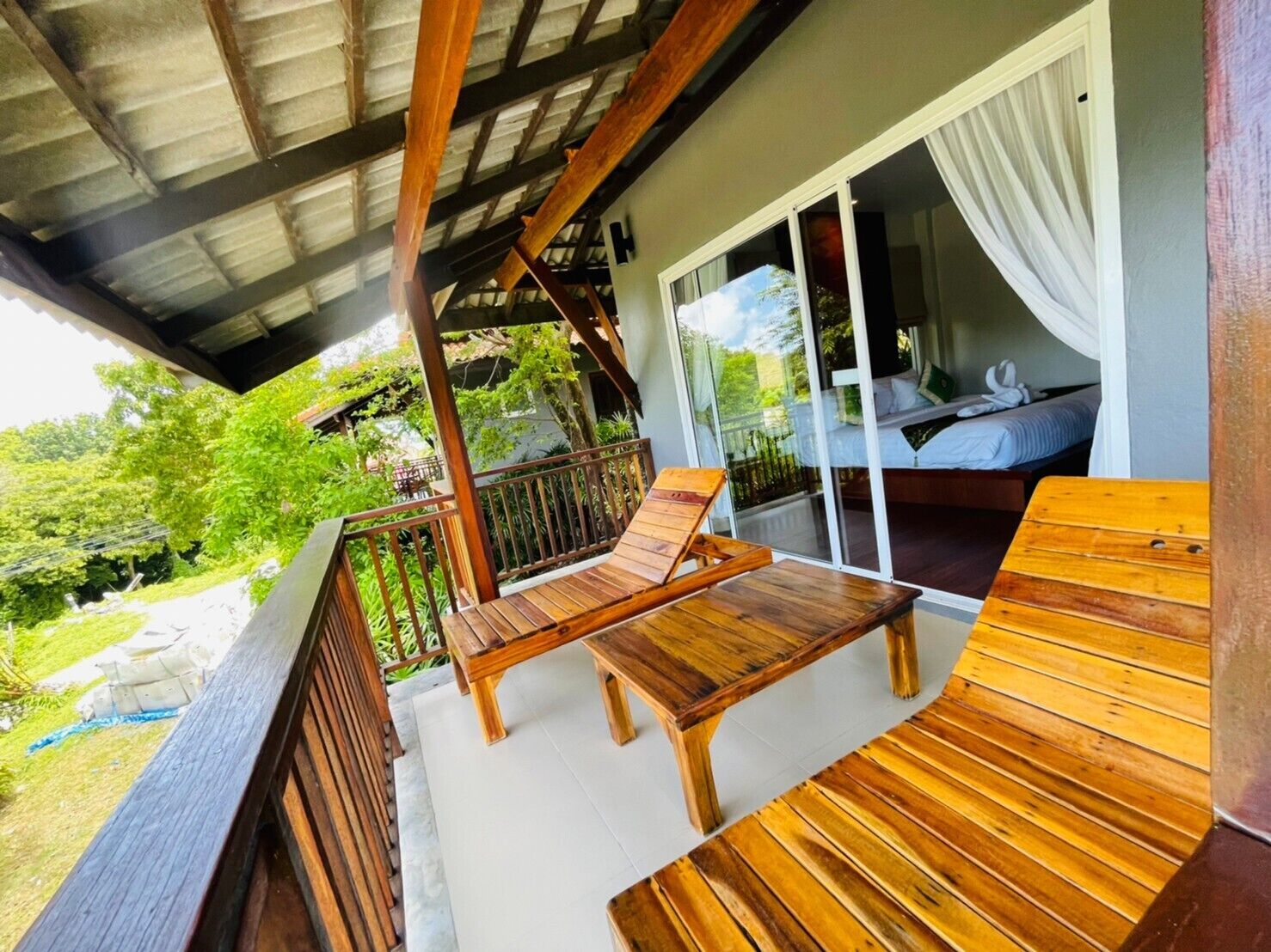 Una terraza de madera con un banco y una mesa, un ventanal de vidrio que da a un dormitorio con cama y cortinas, y vistas a la vegetación exterior.