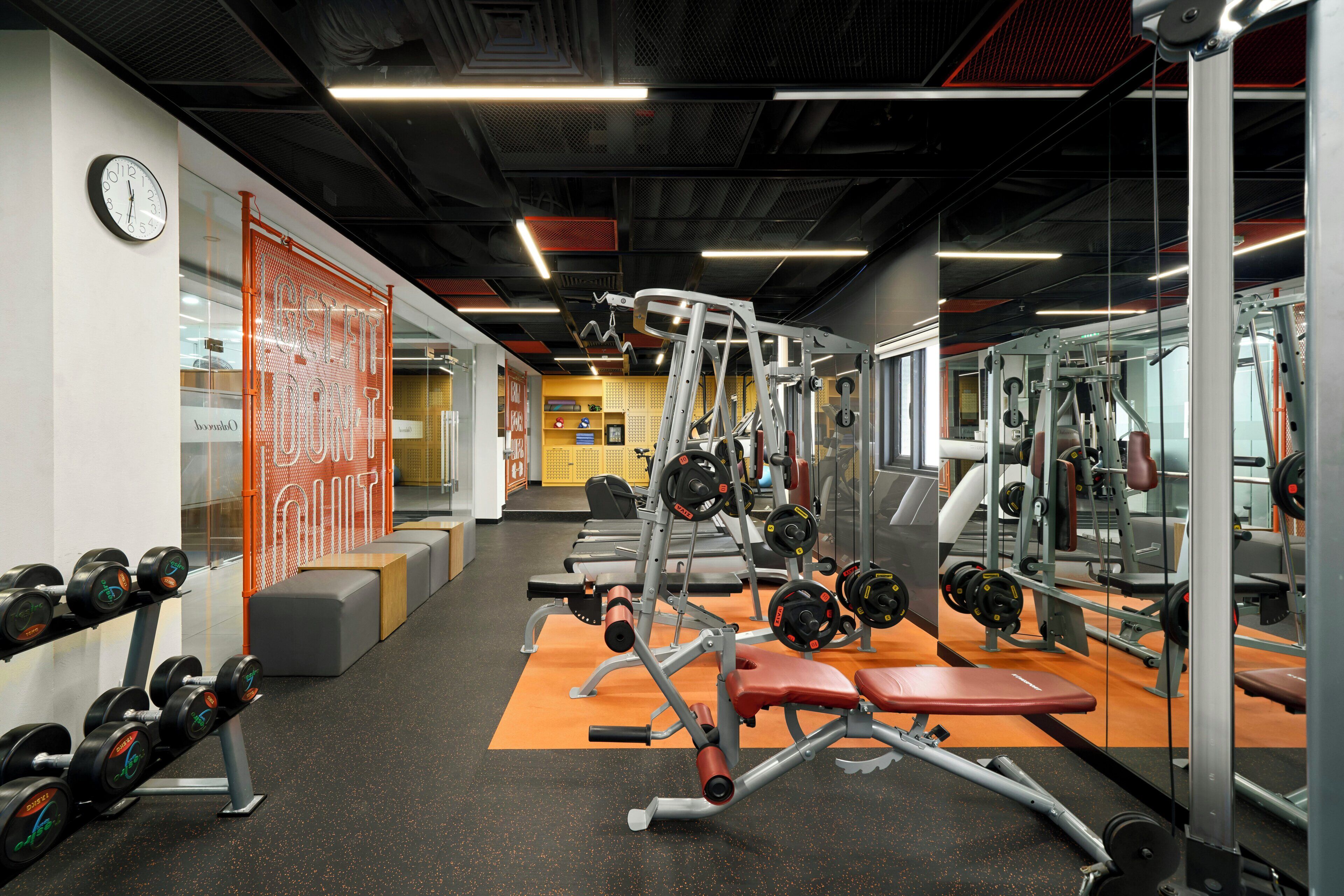 Academia com equipamentos de musculação, bancos e um relógio na parede.