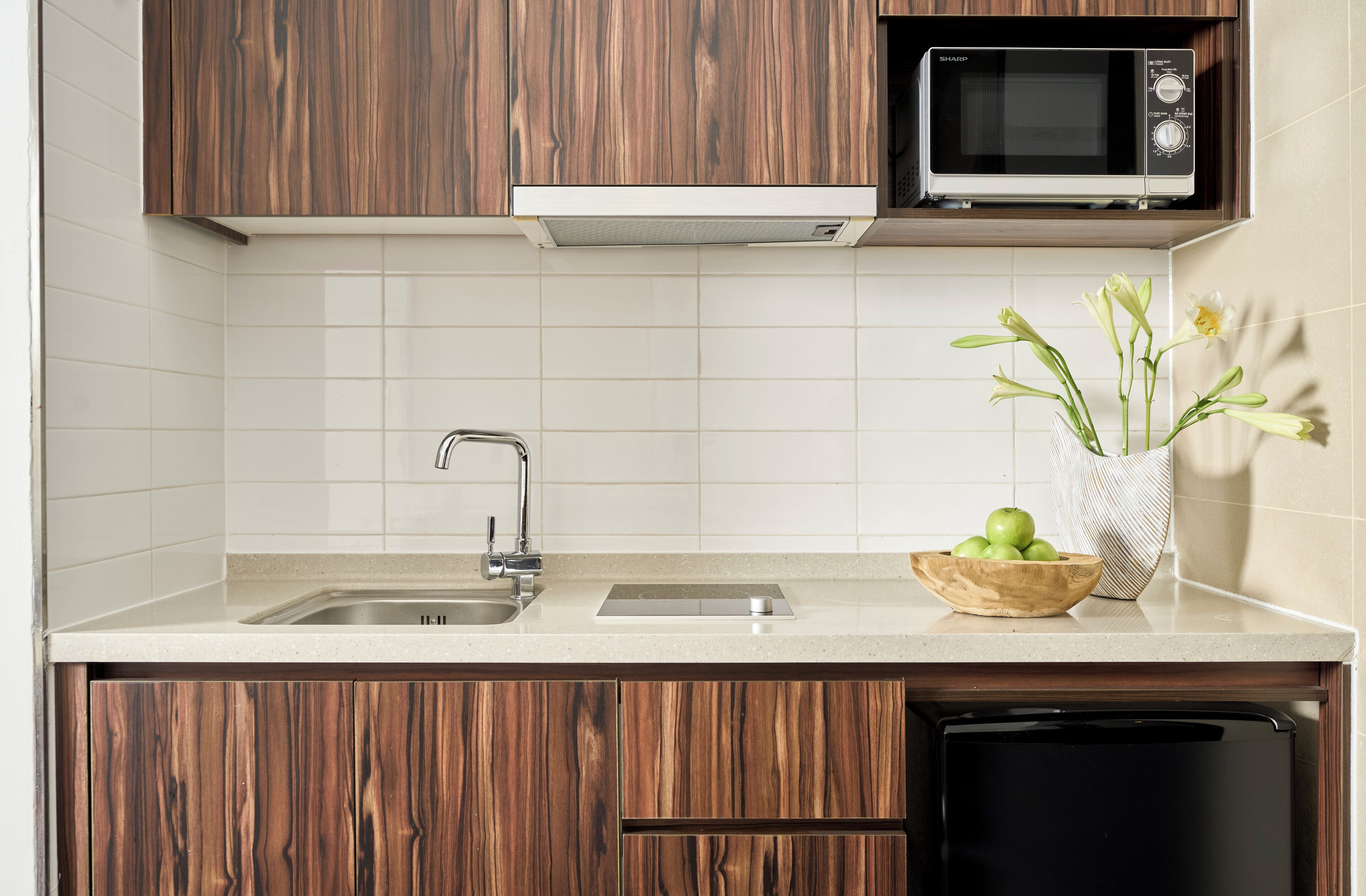 A kitchen with wooden cabinets, a stainless steel sink, a microwave, and a bowl of apples and flowers on the counter.