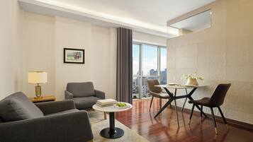 A modern hotel room with a sofa, dining table, and chairs, a city view, and a large window.