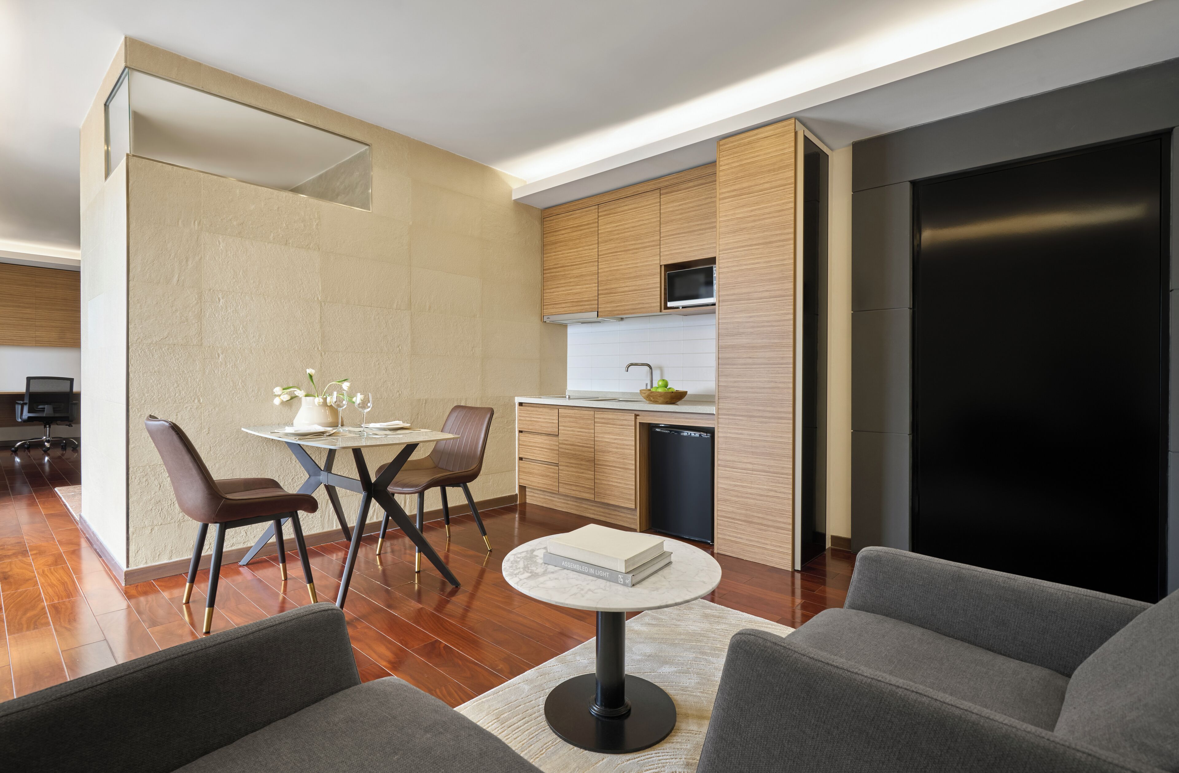 A modern kitchen with wooden cabinets, a built-in microwave, and a dishwasher. There is a dining area with a round table and chairs, and a living area with a sofa and a coffee table.