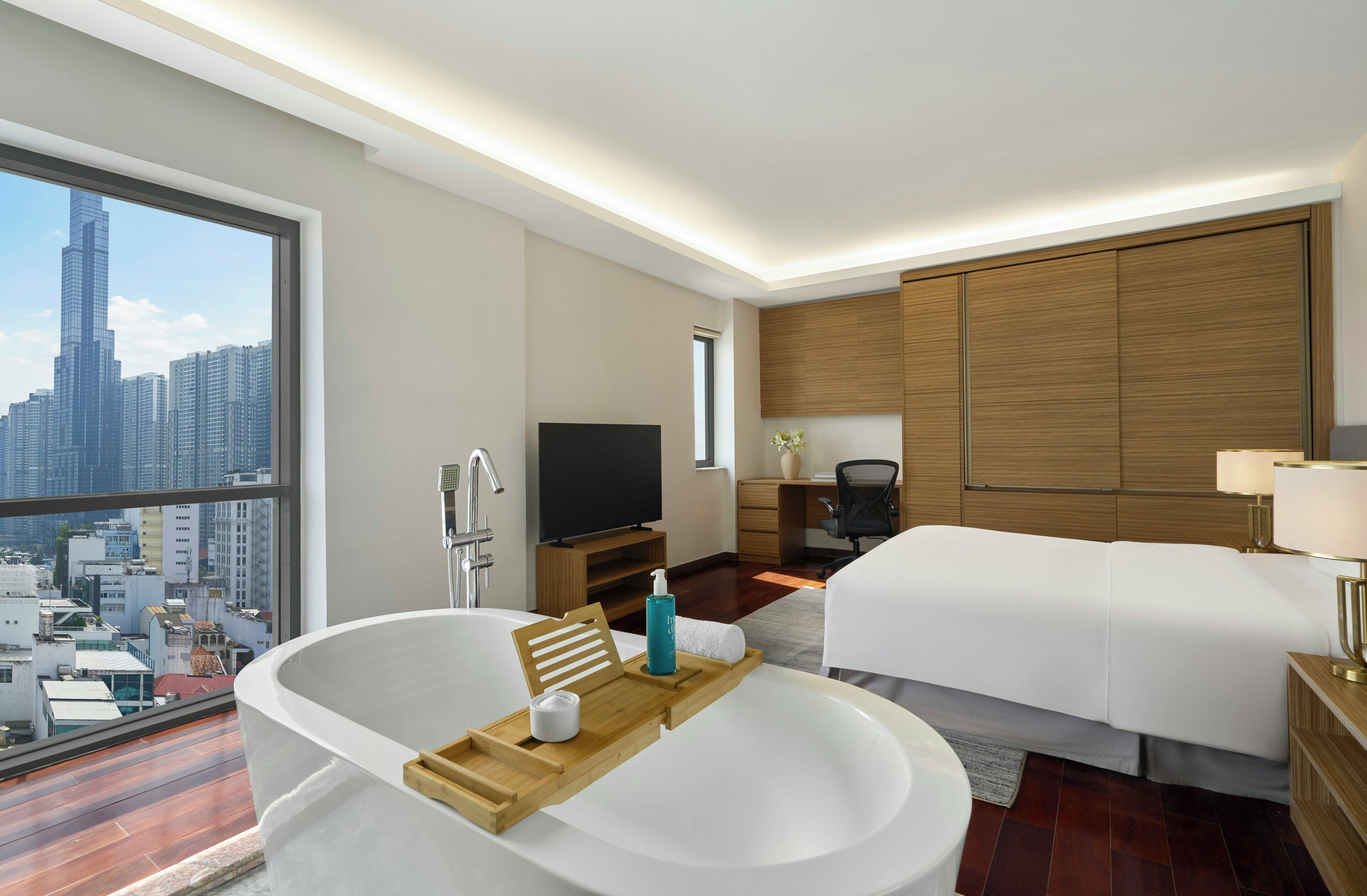 Ein modernes Hotelzimmer mit großem Fenster, einer Badewanne, einem Bett, einem Schreibtisch und einem Fernseher.