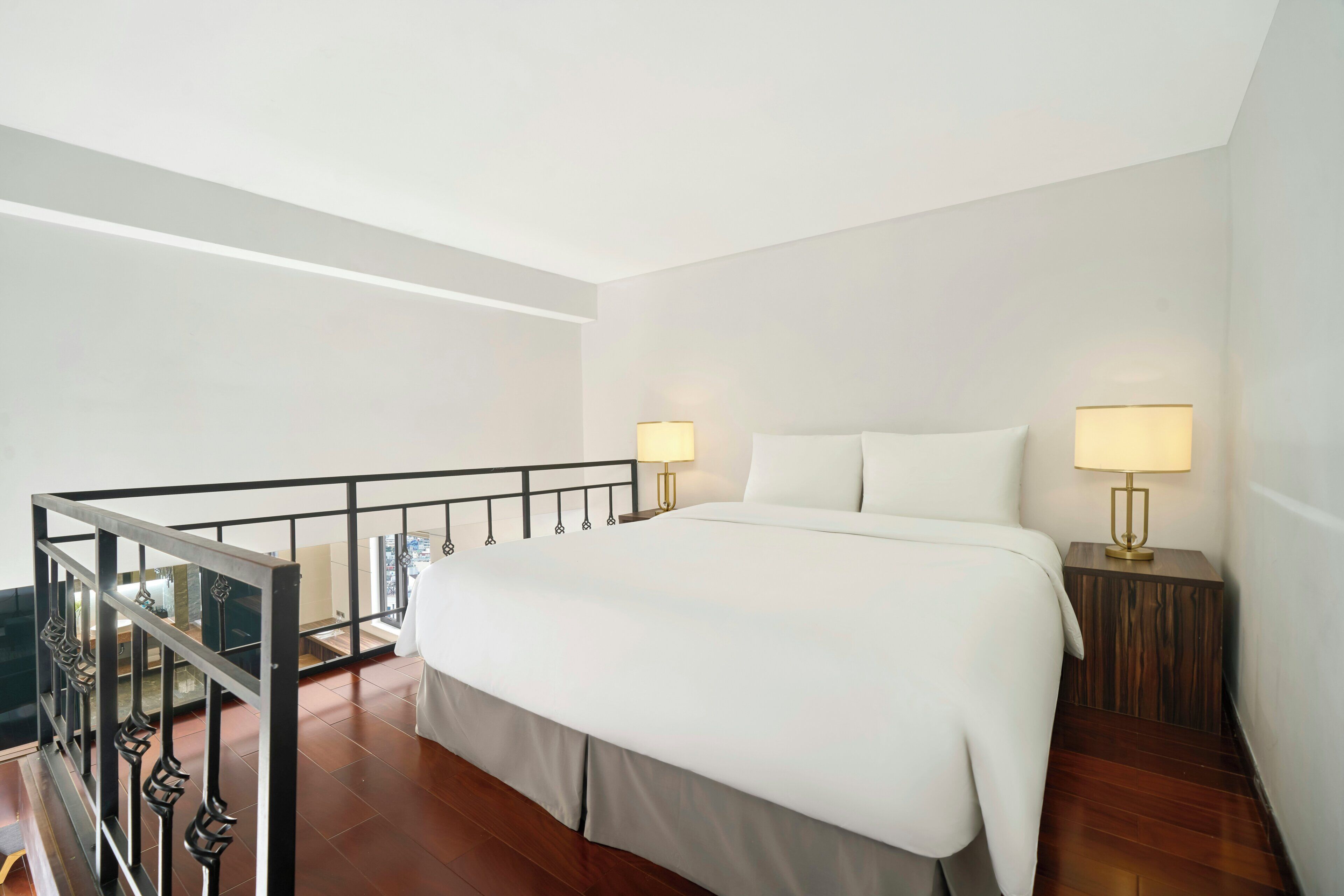 A hotel room with a large bed, two bedside lamps, a wooden nightstand, and a balcony with a railing.
