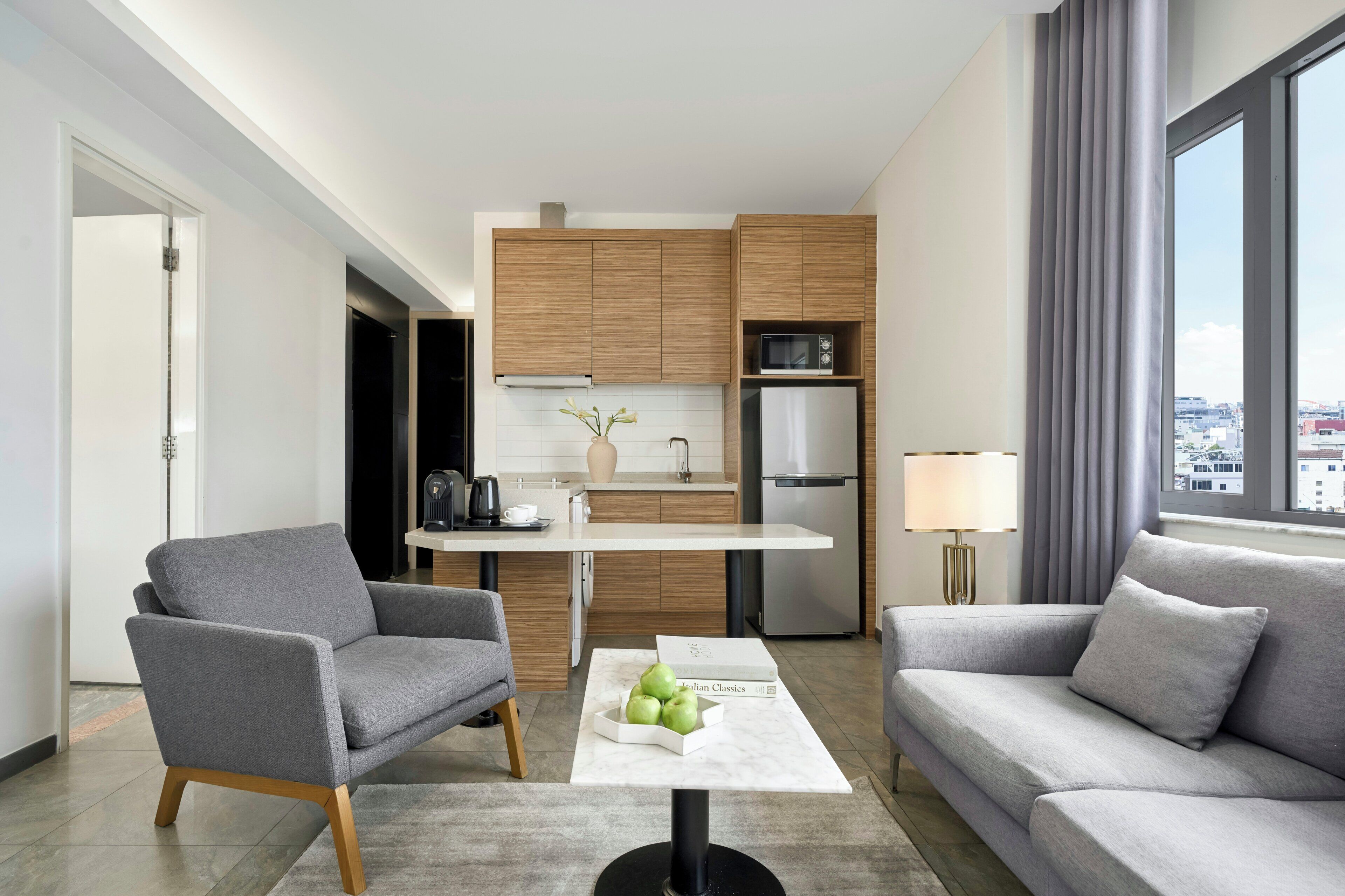 A modern hotel room with a kitchenette, grey sofas, a coffee table with apples, and a city view.