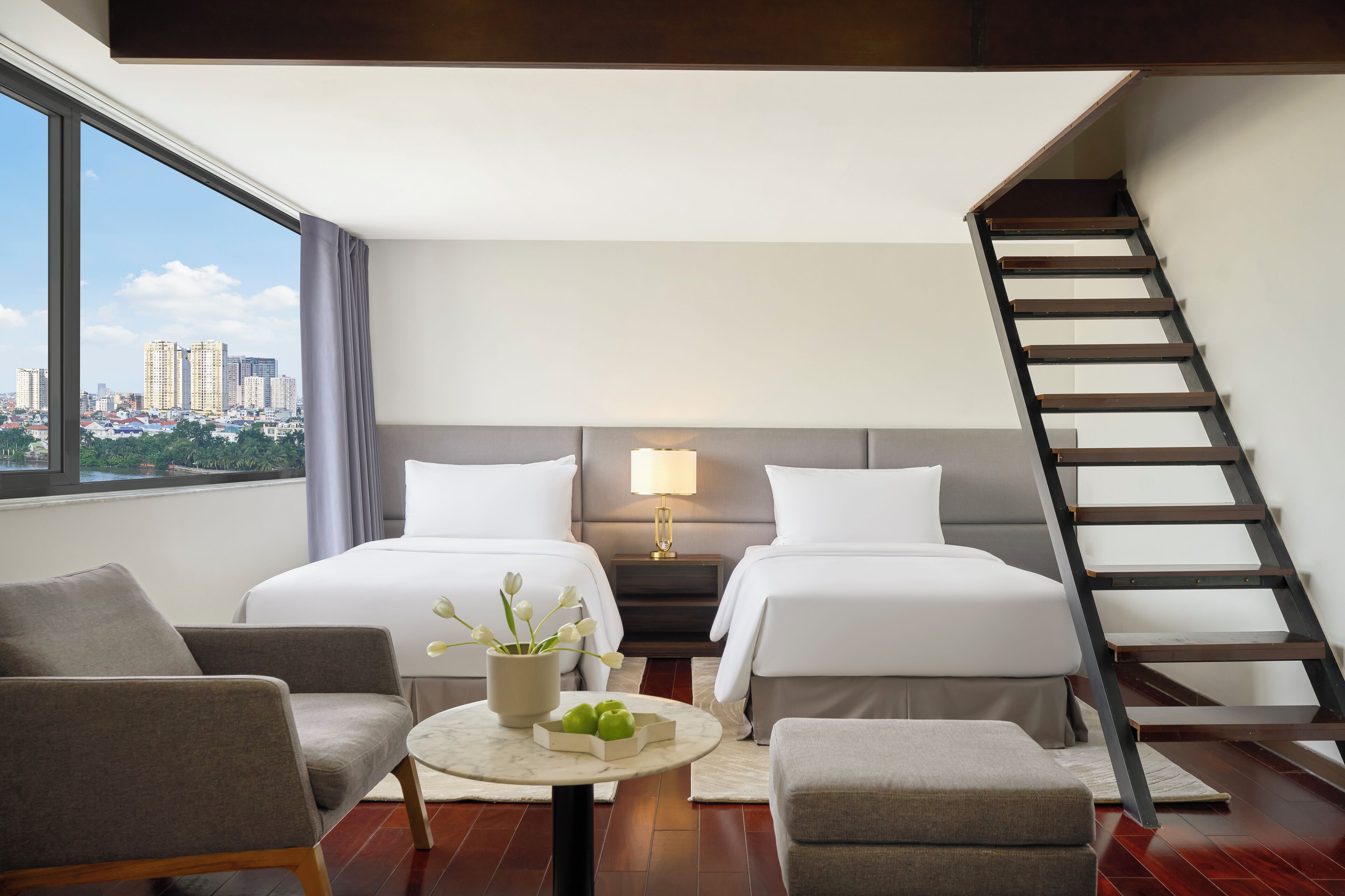 A hotel room with two beds, a sitting area with a table and chairs, a nightstand with a lamp, and a view of the city through large windows.