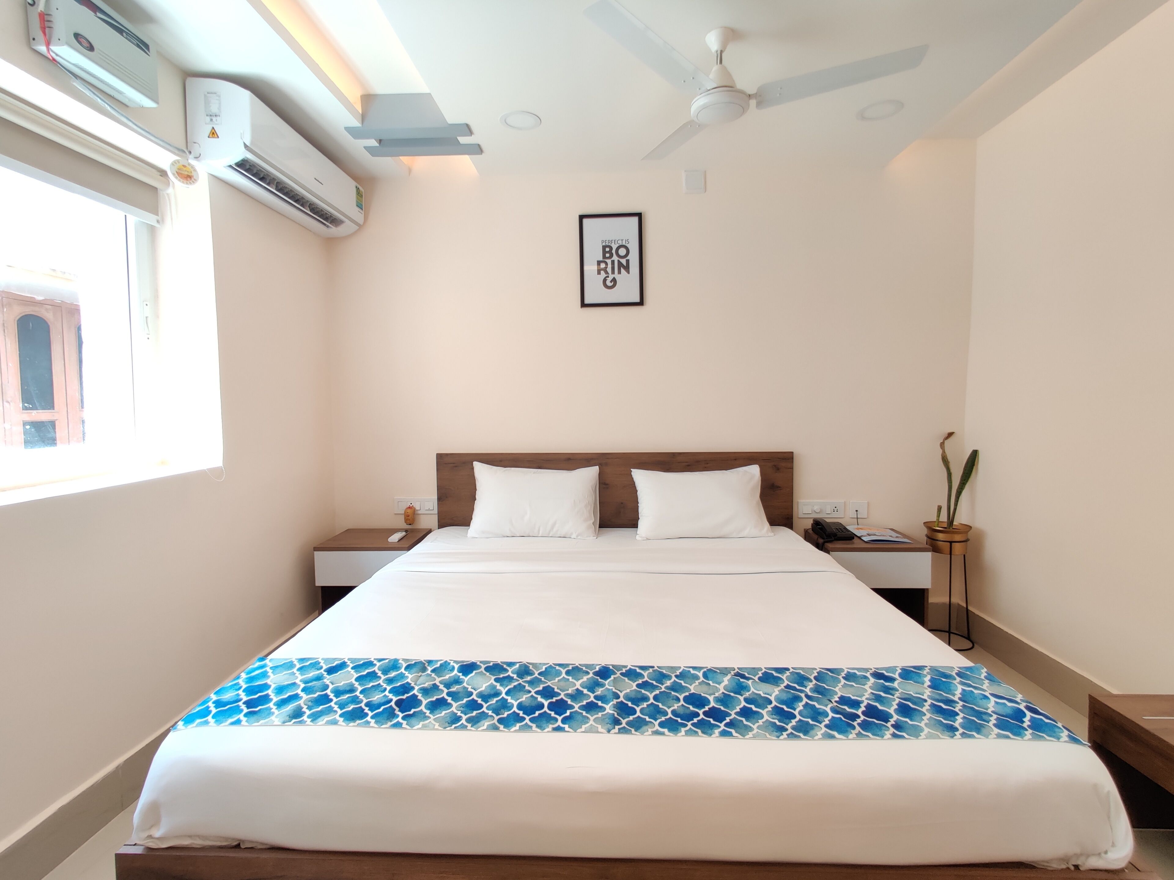 A hotel room with a bed, bedside tables, a ceiling fan, an air conditioning unit, a framed quote on the wall, and a window with a view of a building.