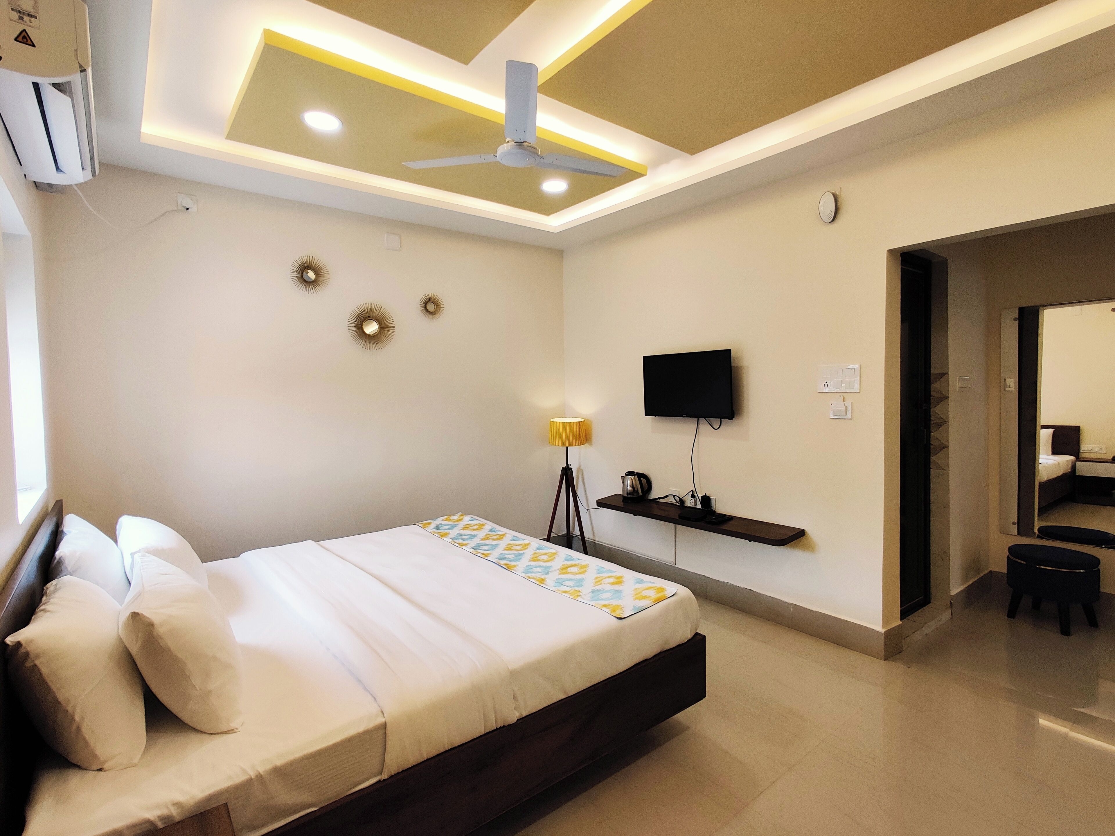 A modern hotel room with a bed, ceiling fan, mounted TV, bedside lamp, and a small shelf with a kettle.