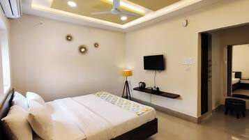 A modern hotel room with a bed, ceiling fan, mounted TV, bedside lamp, and a small shelf with a kettle.