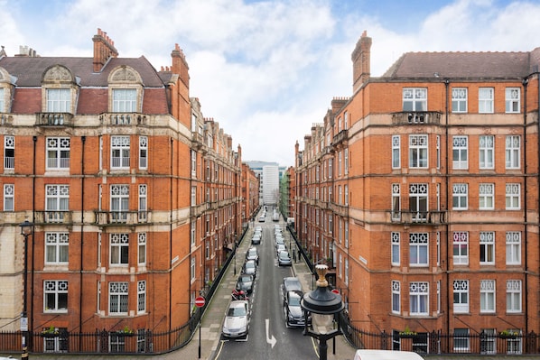 A street view of a row of red brick buildings with uniform windows and balconies, lined with parked cars and a clear sky.