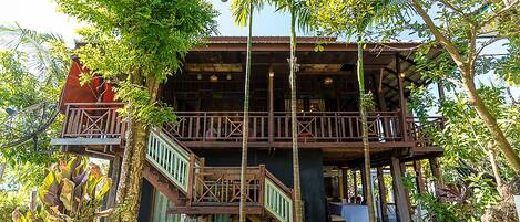 A two-story wooden building with a balcony and stairs, surrounded by lush greenery.