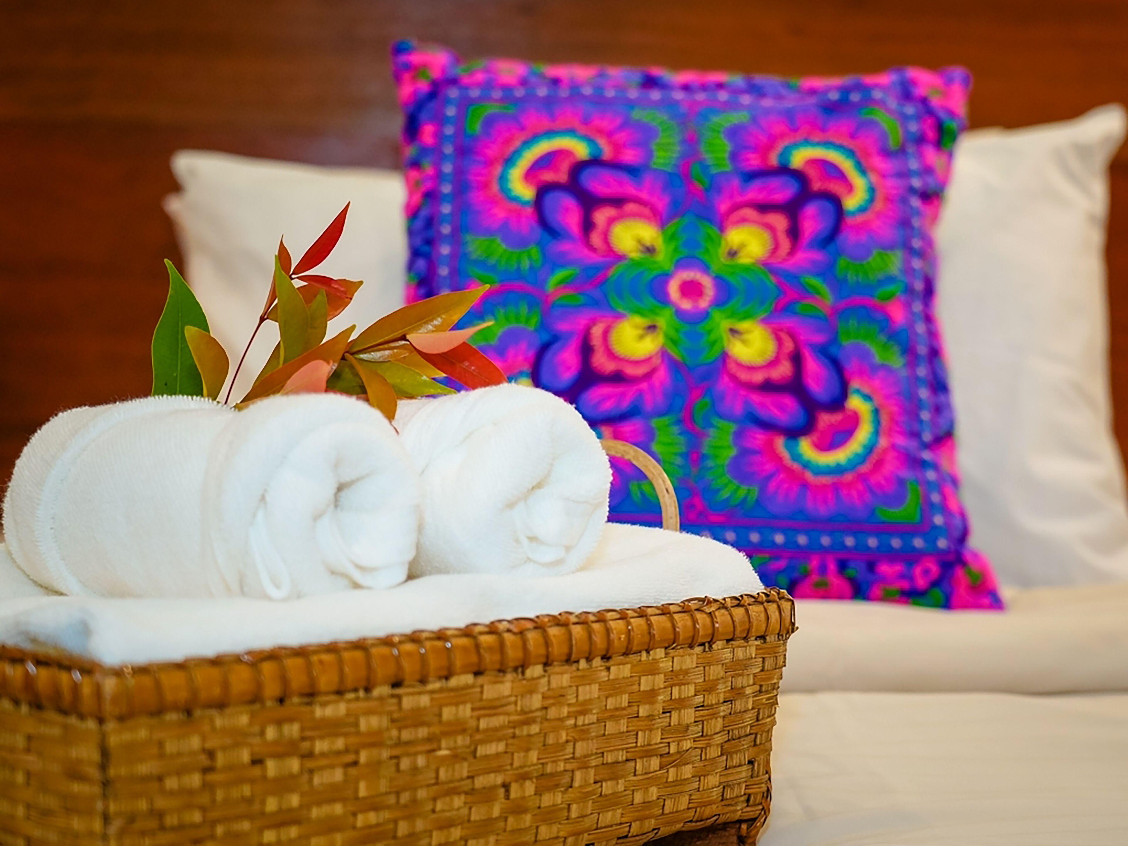 A wicker basket with rolled white towels and decorative leaves on a bed with a colorful pillow.