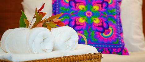 A wicker basket with rolled white towels and decorative leaves on a bed with a colorful pillow.