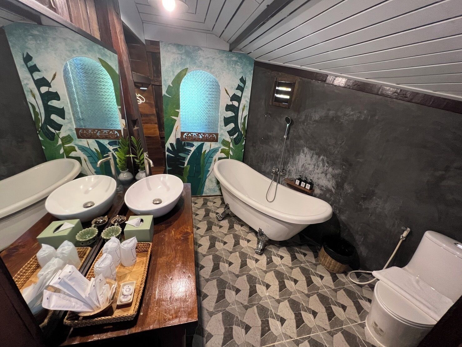 A bathroom with two sinks, a bathtub, and a toilet. There are decorative tiles and a wooden vanity with toiletries.