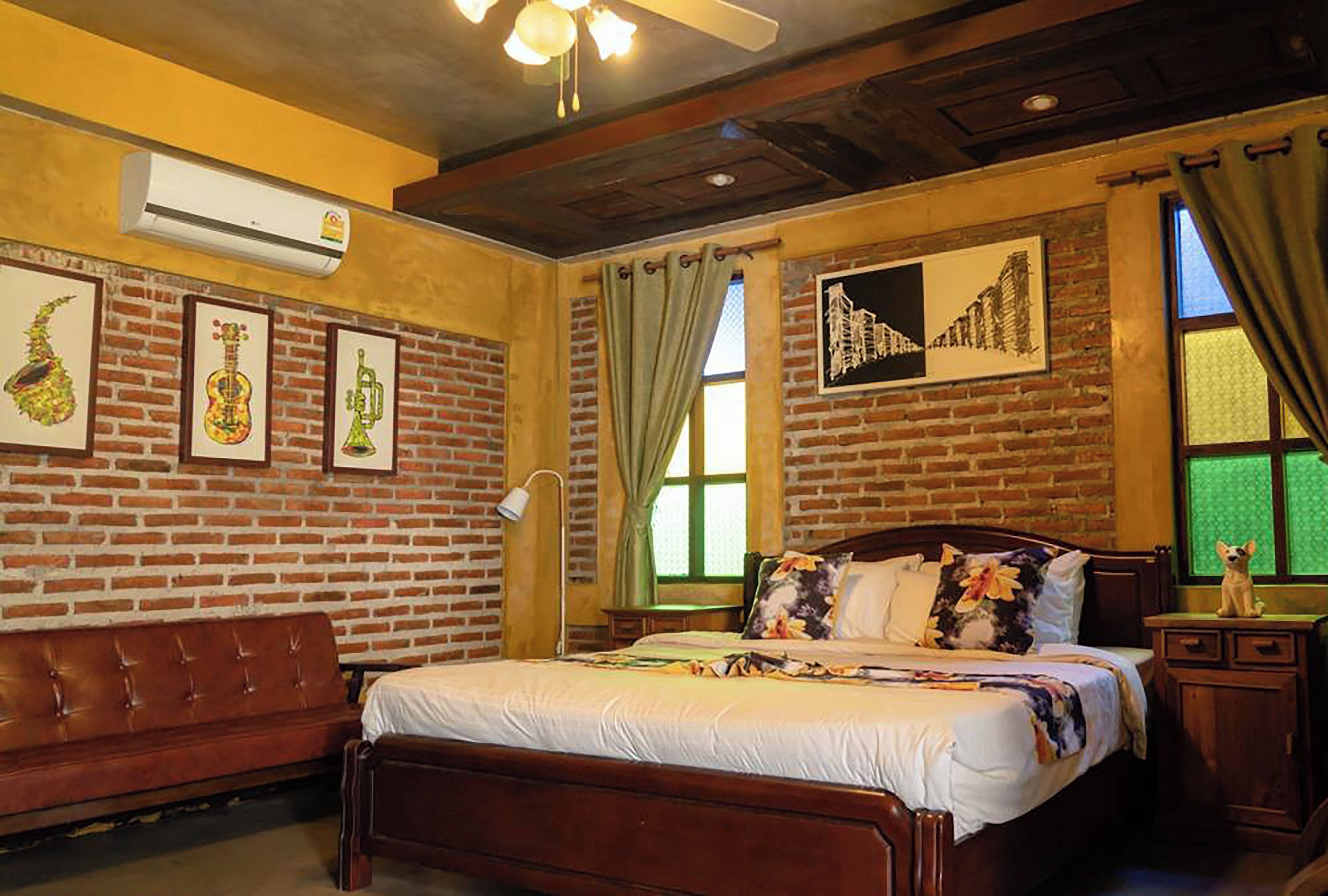A hotel room with a brick wall, a wooden bed, a desk, and a chair. There is a window with curtains and a picture on the wall.